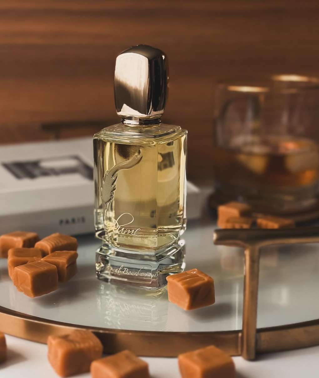 L'image montre un flacon de parfum de la marque Electimuss, mis en valeur par un éclairage coloré. Le flacon a une forme élégante, avec une base rouge et un bouchon doré brillant. À l'arrière-plan, des panneaux de verre colorés dans des tons de jaune, violet et rouge créent un effet de lumière vibrante et réfléchissante. Le design luxueux du flacon, avec un médaillon décoratif, est accentué par les reflets des couleurs, ajoutant une dimension artistique à la composition.