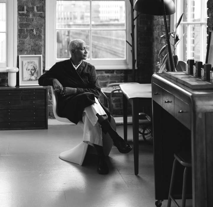 Une femme aux cheveux blonds coupés courts assise dans un fauteuil, regardant par la fenêtre dans un intérieur lumineux avec des murs en briques, une lampe et un meuble en bois.