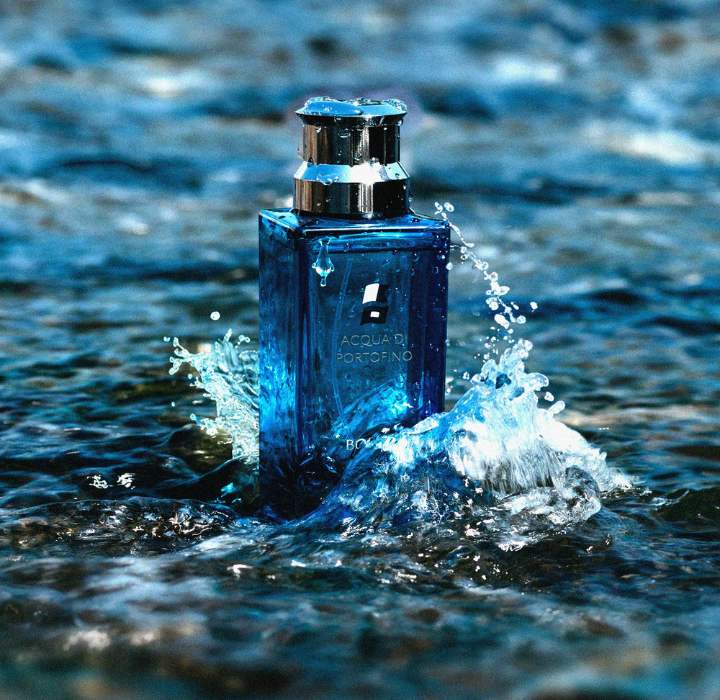 De afbeelding toont een flesje parfum met de naam “ACQUA DI PORTOFINO”, een merk dat wordt verkocht op Scentissime.com, dat uit het water steekt. Het flesje heeft een rechthoekige vorm en is gemaakt van transparant glas met een blauwe tint, wat een gevoel van frisheid en elegantie oproept. De dop van de fles is van metaal, met een glanzende afwerking die het licht vangt. Kleine spatten water stijgen op rond de fles. De achtergrond is wazig, met overvloeiende blauwtinten, wat het idee van een aquatische omgeving versterkt. Deze afbeelding roept een zomerse, mediterrane sfeer op en symboliseert zowel luxe als lichtheid, met een belofte van ontsnapping en frisheid.