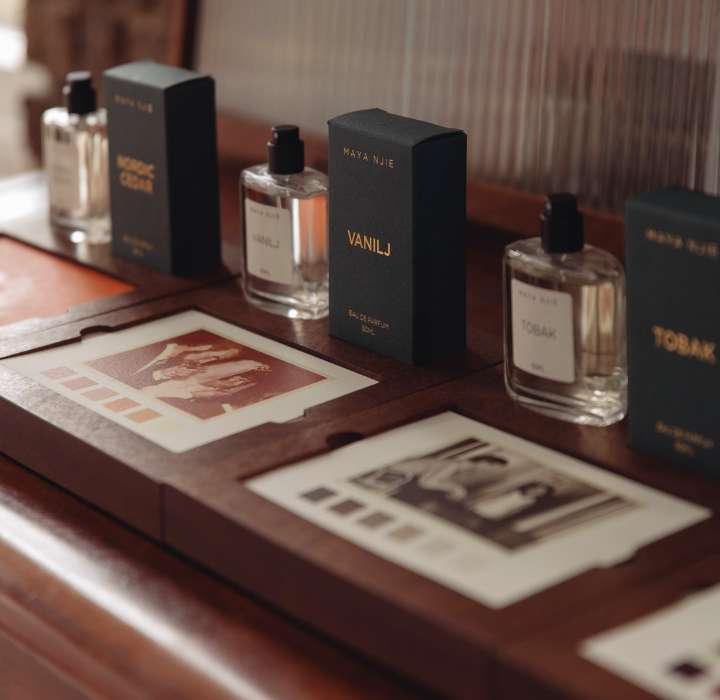 Une collection de flacons de parfum MAYA NJIE posés sur un meuble en bois, avec des photographies en noir et blanc disposées près des flacons.