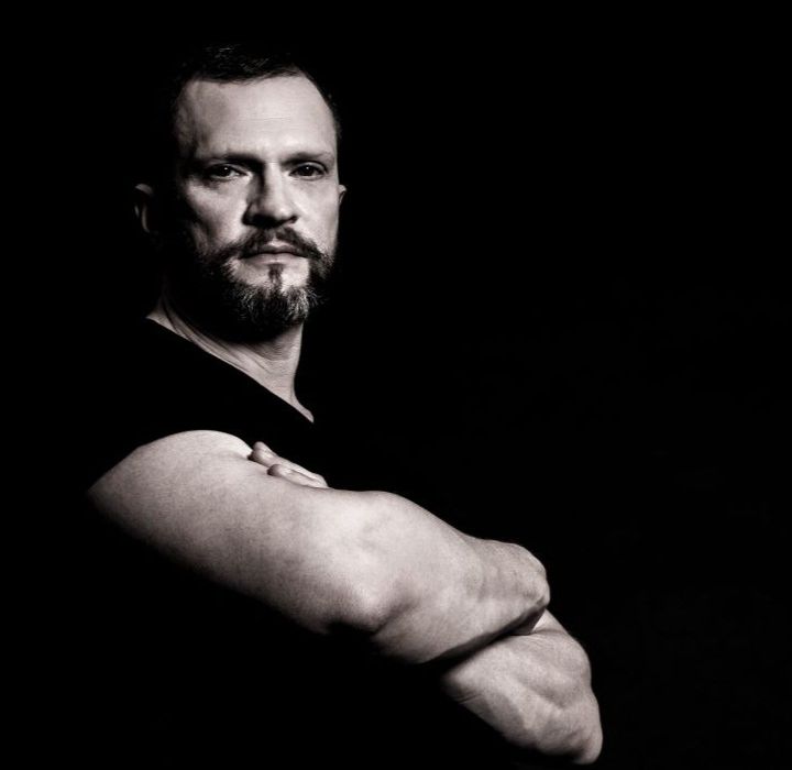 Un homme barbu au regard sérieux, portant un t-shirt noir, se tenant avec les bras croisés contre un fond sombre.