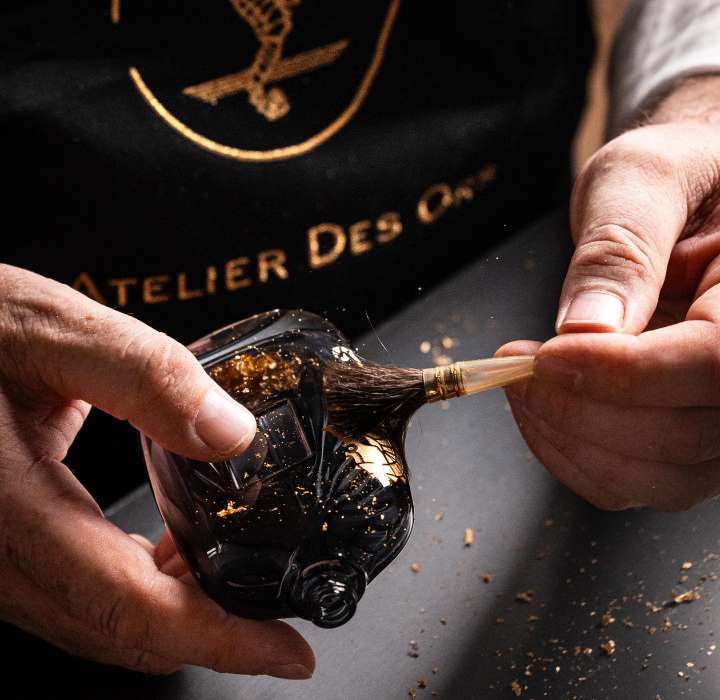 Un homme, portant un tablier, utilise un pinceau pour enlever des particules dorées d'une bouteille en verre noir, entouré de copeaux dorés.