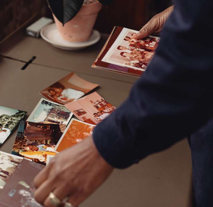 Une main tenant une photo tandis qu'une autre photo est posée sur une table, entourée de plusieurs autres images de souvenirs, avec une plante en pot en arrière-plan.