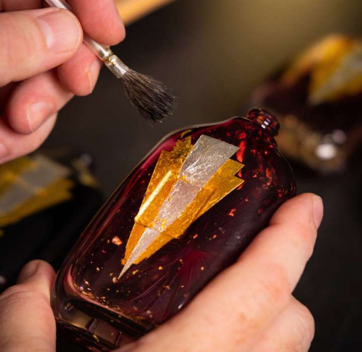 Deux mains tiennent une bouteille en verre rouge, ornée d'un design en or, tandis qu'un pinceau applique des détails dorés sur la surface