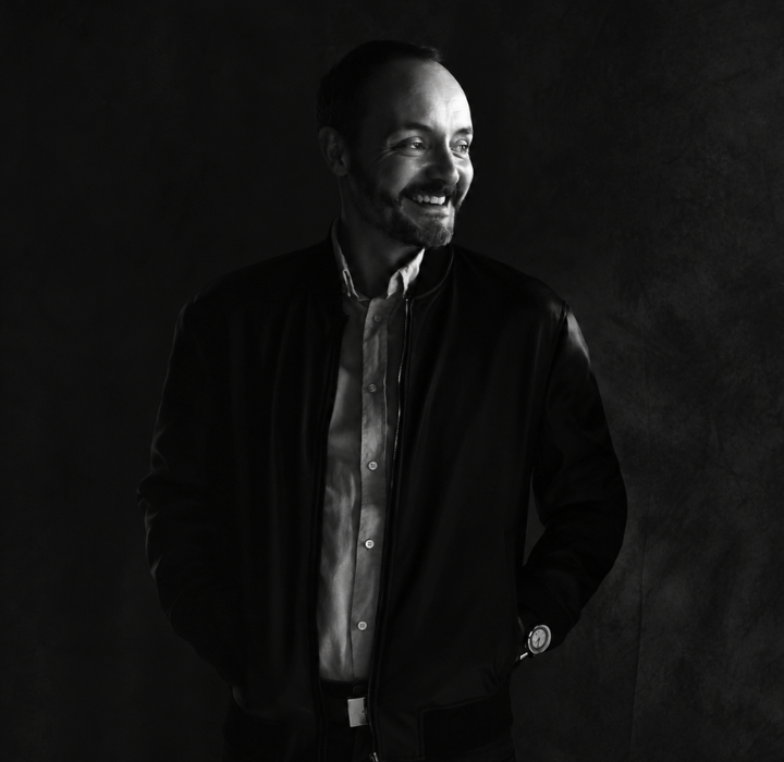 Chiaroscuro portrait of Alexandre Makhloufi, founder of Dissident Parfums. He stands against a black background; only part of his face and one hand are visible. He is smiling.