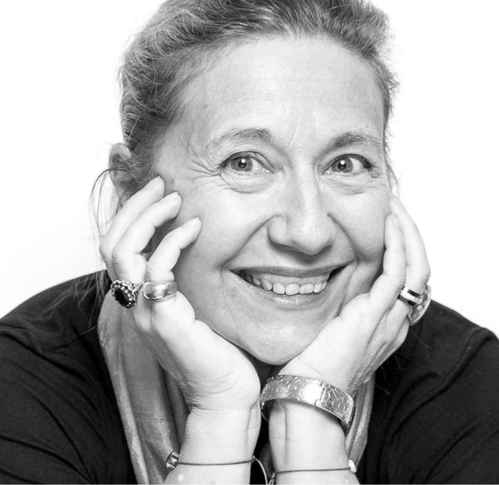 This image features the founder of BINET-PAPILLON, Virginie Choné. We see a smiling woman with light hair and expression lines that reflect her joy. She rests her hands on her face, accentuating her warm smile. She wears several rings and a bracelet, and her background is white, highlighting her expressive and friendly personality.