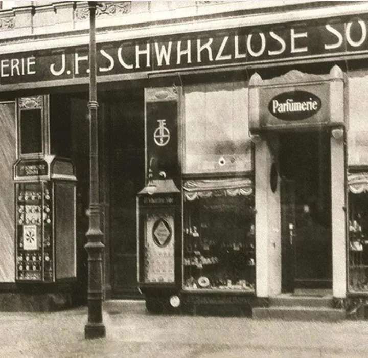 Zwart-witte historische foto van de vintage winkelpui van parfumerie J.F. Schwarzlose Söhne, met art-deco-bewegwijzering, vitrines gevuld met flessen en een straatlantaarn op de voorgrond.