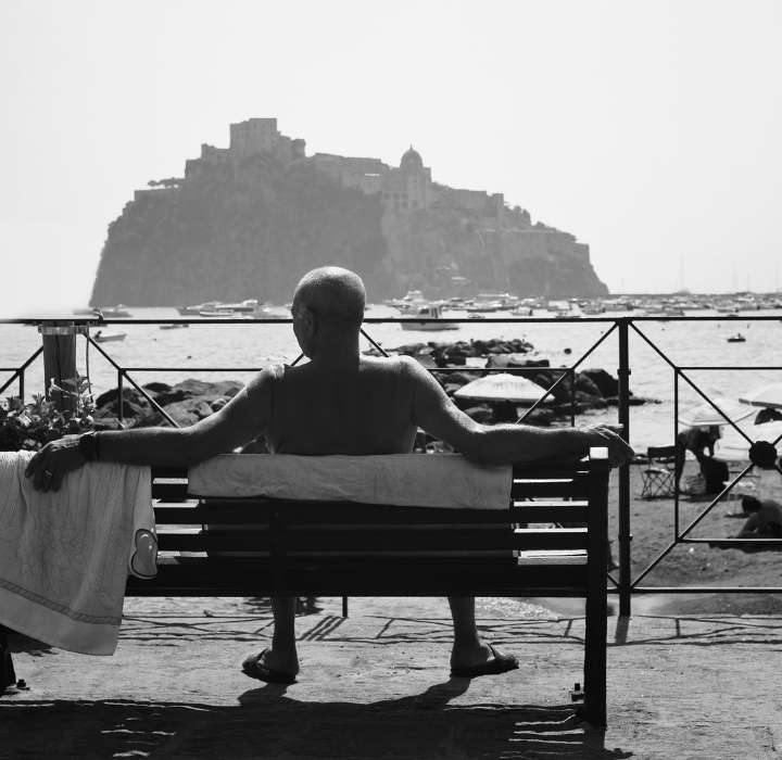 Una fotografia in bianco e nero di un uomo a torso nudo seduto su una panchina di legno, con le braccia aperte, rivolto verso il mare. In lontananza, si vedono delle barche nel porto e una grande fortezza su una collina, sotto un cielo limpido.
