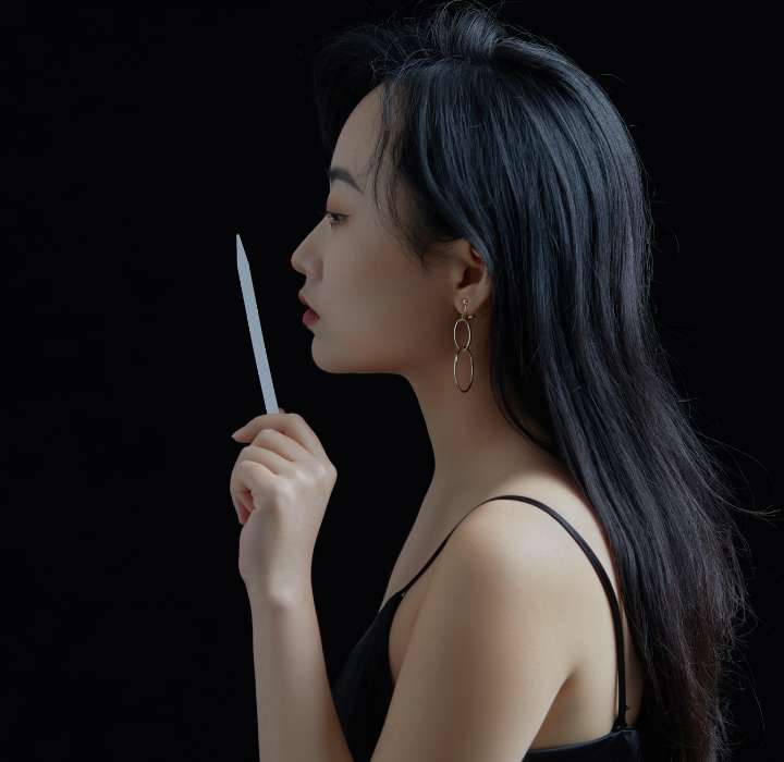 L'image montre une femme vue de profil, avec des cheveux longs et noirs qui tombent doucement sur son épaule. Il s’agit de Lam Xiao, fondatrice de la marque de parfum de niche TOMBSTONE, vendue sur Scentissime. Elle est vêtue d'un haut noir sans manches, ce qui accentue la simplicité et l'élégance de sa silhouette. Elle tient délicatement une touche à sentir près de ses lèvres. Ses yeux sont fermés, et son expression est calme et contemplative. Elle porte des boucles d'oreilles dorées qui ajoutent une touche de brillance à sa tenue. L'arrière-plan est noir, mettant en valeur ses traits et l'objet qu'elle tient. L'ensemble de la composition évoque une ambiance paisible et introspective.