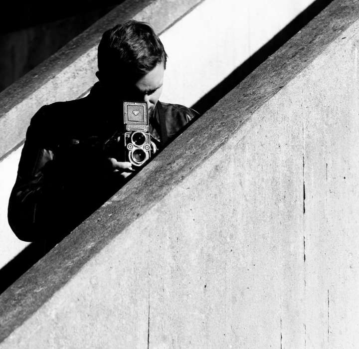 Foto in bianco e nero: un uomo dai capelli scuri sta scattando un selfie con una vecchia macchina fotografica e una seconda fotocamera. Indossa una giacca scura e si riflette su una parete chiara, con una scalinata sullo sfondo.