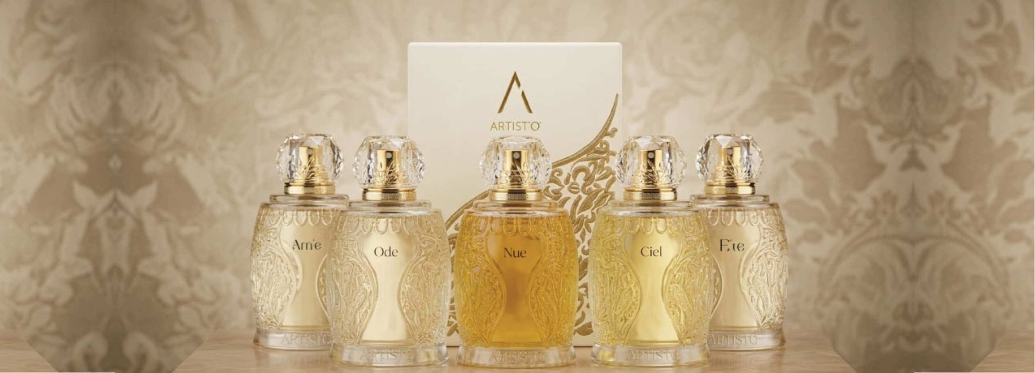 Foto von sechs Parfümflakons aus Glas, deren Farbe von hell bis bernsteinfarben reicht, vor einem beigen, mit Motiven verzierten Hintergrund. Die Verschlüsse sind transparent und haben einen goldenen Sockel. Auf den Etiketten sind unter anderem die Aufschriften „Ame“, „Ode“, „Nue“, „Ciel“ und „éte“ zu lesen. In der Mitte befindet sich eine weiße Schatulle mit dem Logo „ARTISTO“.