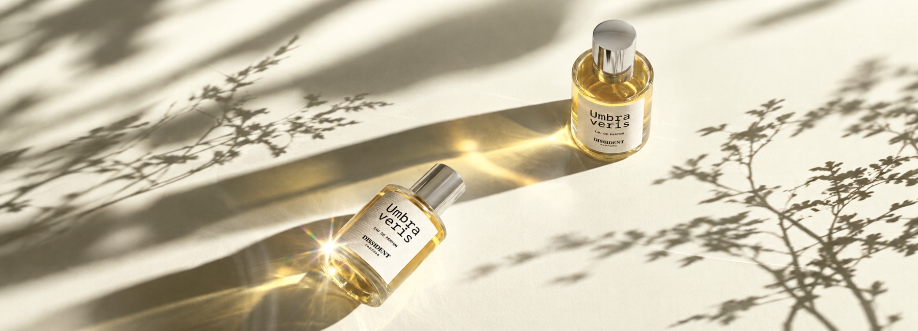 Two bottles of Umbra Veritas perfume by the brand Dissidents Parfums are placed on a light-colored surface. Their shadows cast a long silhouette. Shadows of vegetation can be seen around the bottles.
