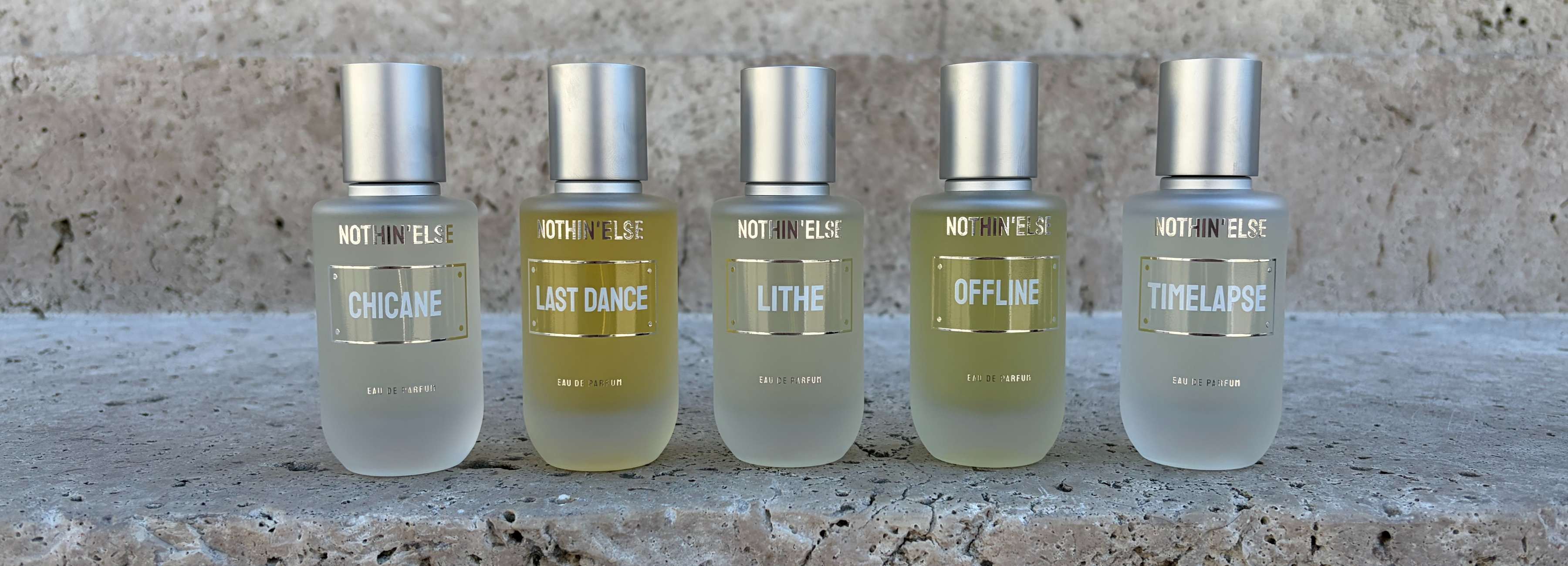 Una fotografia di cinque flaconi di profumo allineati su una superficie di pietra. Ognuno ha un tappo d'argento e un'etichetta rettangolare. I nomi 'CHICANE', 'LAST DANCE', 'LITHE', 'OFFLINE' e 'TIME LAPSE' sono visibili sui flaconi. Il testo "NOTHIN' ELSE" è visibile sopra ogni etichetta.