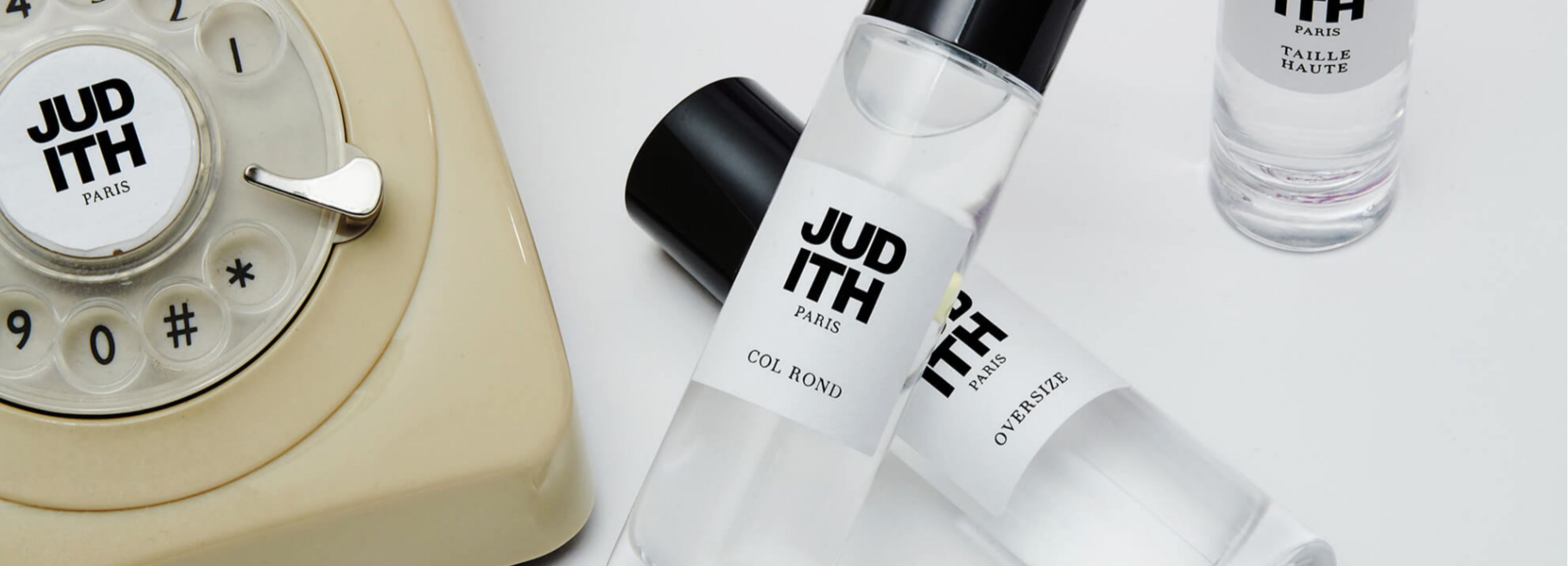 Una composizione ordinata di diverse bottiglie di profumo JUDITH Paris, con "COL ROND" e altre etichette visibili, e un telefono antico con un quadrante rotante color crema sulla sinistra dell'immagine.