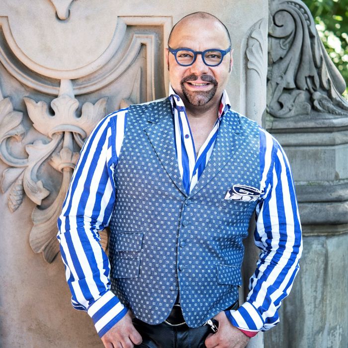 Il profumiere Rodrigo Florès-Roux si trova davanti a una facciata ornata. Indossa un abbigliamento dai colori vivaci, una camicia a larghe righe bianche e blu e un gilet blu decorato con motivi bianchi.