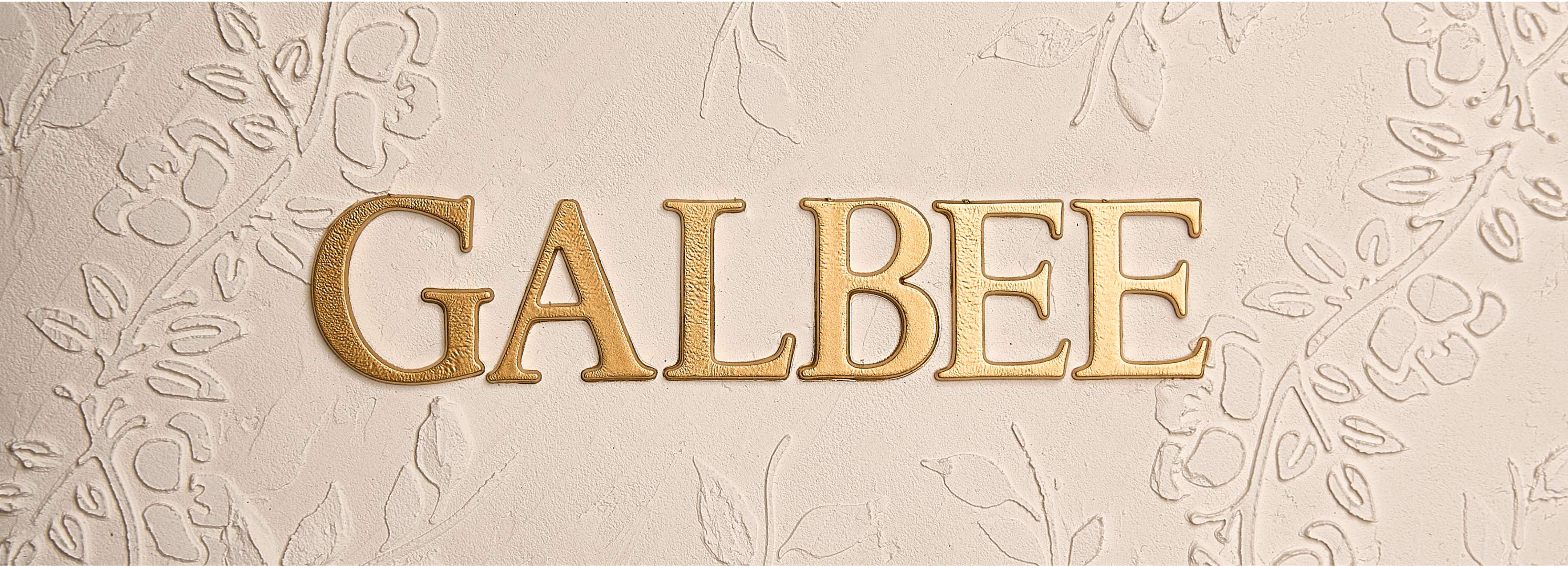 Op de afbeelding staat het woord “GALBEE” in gouden blokletters. De letters hebben een glanzende afwerking die in het oog springt en een indruk van luxe geeft. De achtergrond heeft structuur, waarschijnlijk gips of stucwerk, met een subtiel patroon van bladeren en bloemen, wat een interessant contrast vormt met de gouden letters. De achtergrondkleur is licht, wat de naam benadrukt.