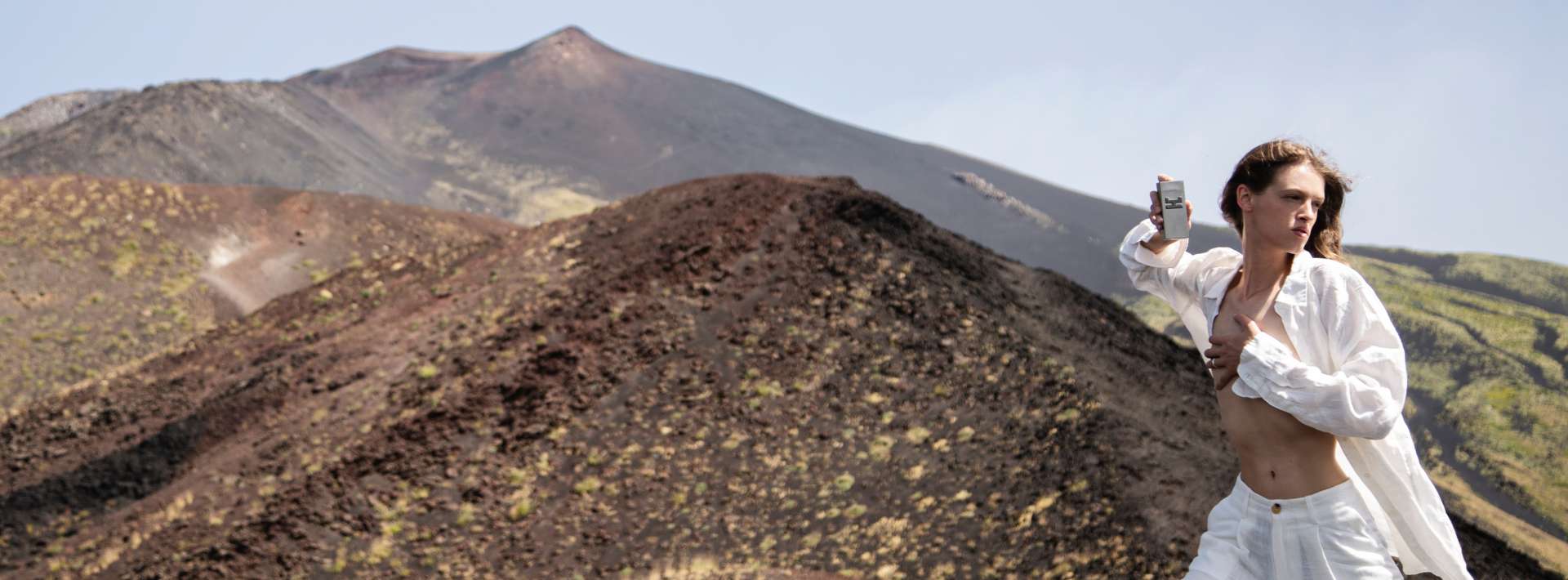 The image shows a man with long hair posing in a volcanic landscape. He is wearing an open white shirt that reveals his midsection, and light-colored pants. He is standing, turned slightly to the side, and holding a perfume bottle at chest height, as if presenting or brandishing it. In the background, volcanic hills are visible, with shades of black, brown, and green, suggesting a rich but arid land. The sky is clear, adding brightness to the scene. The ensemble creates a dynamic and bold atmosphere, blending the beauty of nature with a sense of fashion and attitude.