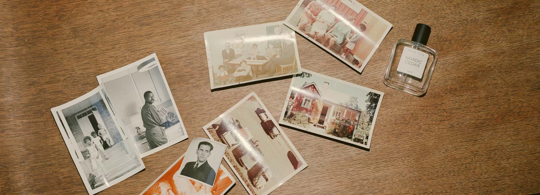 Une collection de vieilles photographies en noir et blanc et en couleur disposées sur une table en bois, avec un flacon de parfum moderne de la marque MAYA NJIEà côté.