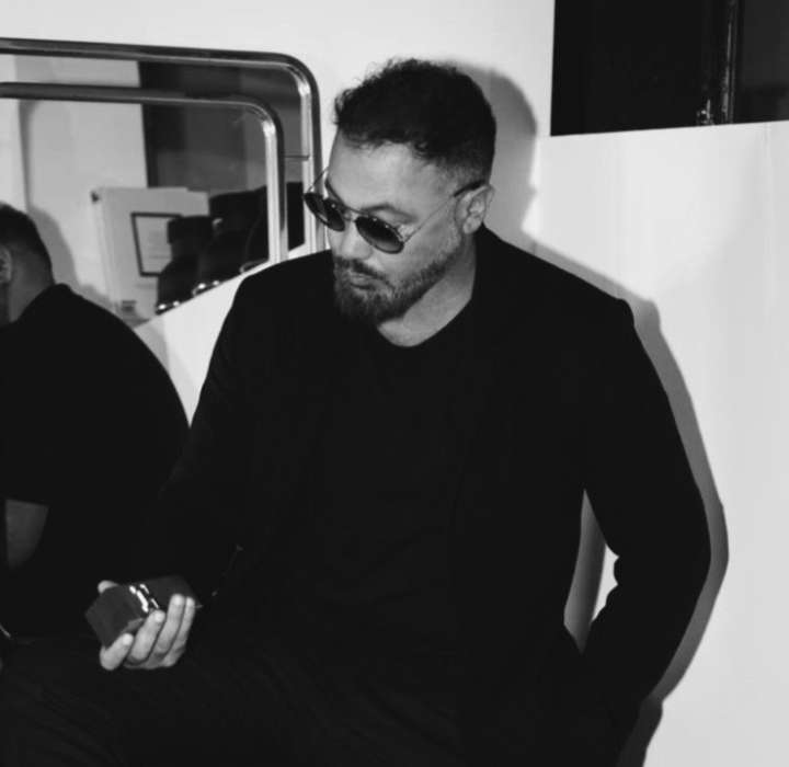 The image shows a seated man wearing a black suit that gives him an elegant and sophisticated look. This man is Cael Pipin, founder of the HEMCAEL brand. He wears dark sunglasses that add a touch of mystery to his expression. The man has a well-trimmed beard and a thoughtful gaze, concentrating on a perfume bottle he holds in one hand. The background is blurred, but modern decor elements can be seen, such as reflective surfaces that create a play of light. The lighting is soft, reinforcing the chic and contemporary atmosphere of the scene. The overall effect exudes a sense of confidence and style.