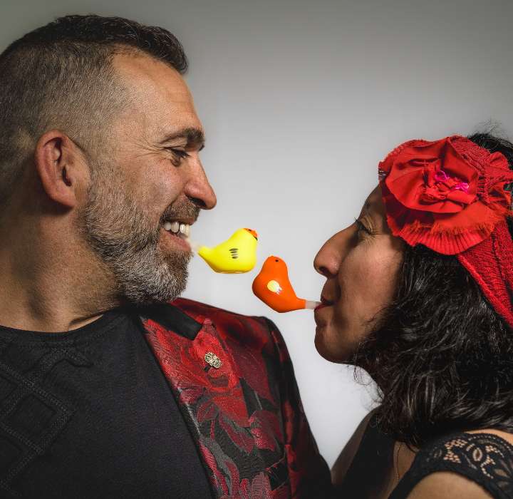 De afbeelding toont een man en een vrouw die tegenover elkaar staan en naar elkaar glimlachen. De man draagt een rode blazer met bloemenpatronen en heeft een grijze baard. De vrouw draagt een rood haarband en heeft donker haar. Tussen hen staan twee kleine plastic vogels, een gele en een oranje, die met hun snavels elkaar lijken te raken. De achtergrond is neutraal, waardoor hun vrolijke interactie opvalt.