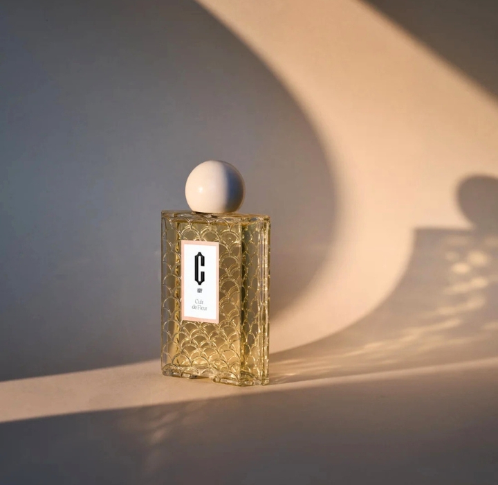 A bottle of Cray perfume is placed in front of a curved backdrop, with late-day light casting a curved shadow, creating a lace-like effect from the bottle’s decoration.