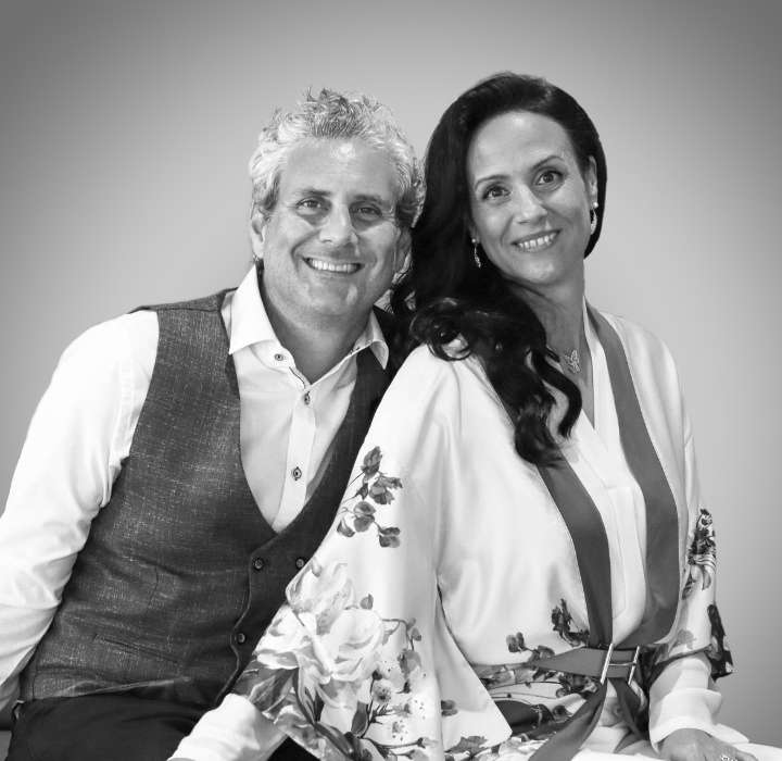 The image shows the founding couple of the fragrance brand PLUME IMPRESSION. They are sitting next to each other, smiling. They are in black and white. The man is wearing a white shirt under a grey waistcoat, showing off his casual but well-groomed style. His hair is grey and curly, and his expression is warm and welcoming. The woman on his right is wearing a flowing floral dress. Her hair is long and wavy, and she has a friendly smile on her face. She exudes an aura of confidence and grace. The whole composition conveys a feeling of complicity and conviviality between them, suggesting a friendly or romantic relationship.