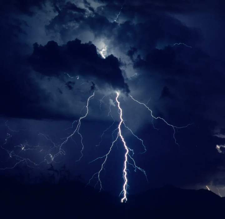 Das Bild zeigt einen stürmischen Himmel, der mit dunklen, bedrohlichen Wolken gefüllt ist. In der Mitte des Bildes sticht ein kraftvoller Blitz hervor, der von den Wolken zum Boden herab schlägt. Das Licht des Blitzes ist sehr intensiv und beleuchtet die Szene kurzzeitig mit einem strahlend weißen Glanz.

Die Wolken sind in einem sehr dunklen Grau, fast Schwarz, und wirken wirbelnd, was das Gefühl von Spannung und Sturm verstärkt. Unten sind Silhouetten von Bergen sichtbar, die einen Kontrast zum hellen Licht des Blitzes bieten.