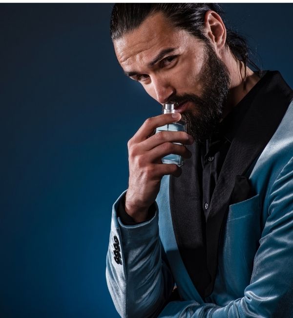 Un homme barbu aux cheveux noirs, habillé élégamment, sent le col d'un flacon de parfum