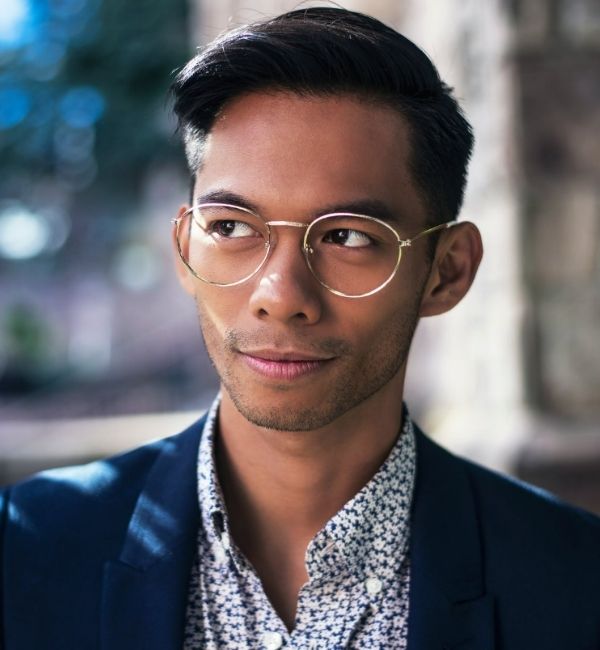 Un homme élégant, porte des lunettes rondes cerclées de métal, une chemise à motifs et une veste bleue.