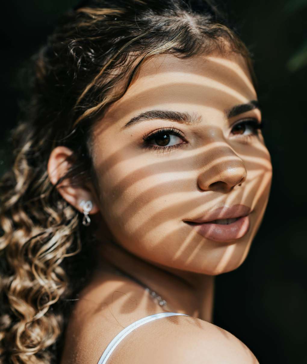 The image is a portrait of a young woman, looking over her shoulder with a slight smile. Her face is partially shaded by horizontal lines of light, as if she were standing in the shadow of a slatted structure or foliage.

Her hair is curly and appears to have lighter strands, framing her face. She is wearing a thin garment strap, suggesting a light outfit.

The background is dark and out of focus, emphasizing the woman's face and creating a striking contrast with the lit areas.