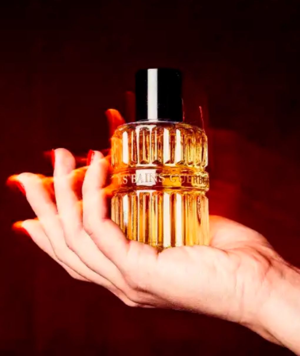 Flacon de parfum Les Bains Guerbois présenté dans une main aux ongles rouges sur fond rouge sombre