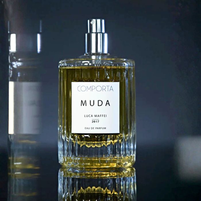 Een transparant glazen parfumflesje, rechthoekig van vorm met facetten, met daarin een gouden vloeistof. Op het witte etiket staat "COMPORTA MUDA, LUCA MAFFEI, 2017, Eau de Parfum".