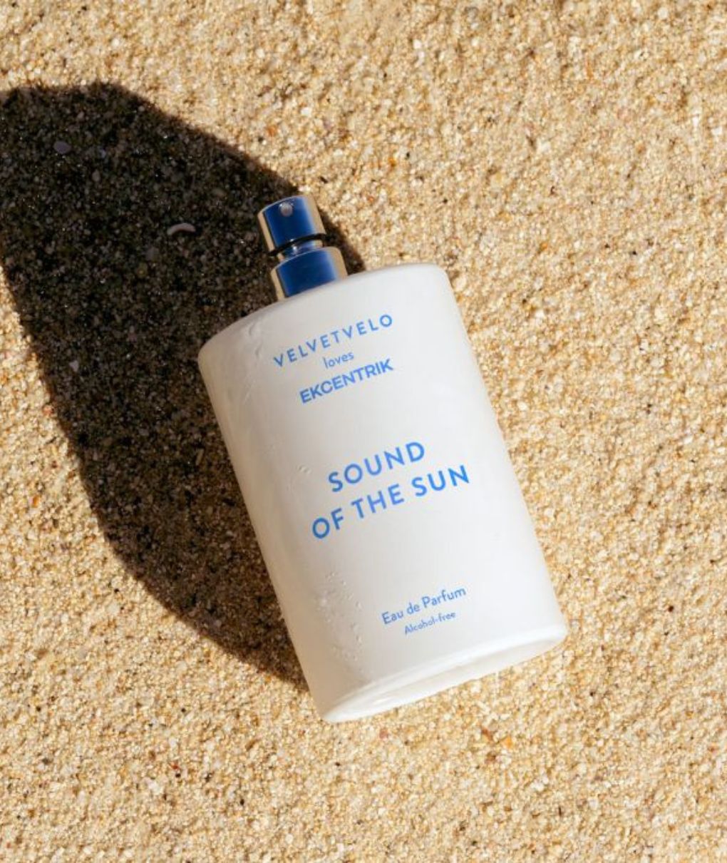 L'image présente un flacon de parfum blanc posé sur du sable. Le flacon est cylindrique avec un vaporisateur argenté sur le dessus. Sur l'étiquette, on peut lire en bleu les inscriptions "VELVETVELO" et "SOUND OF THE SUN". Le sable environnant est de couleur beige clair. L'ombre du flacon est visible sur le sable, indiquant une source de lumière vive.