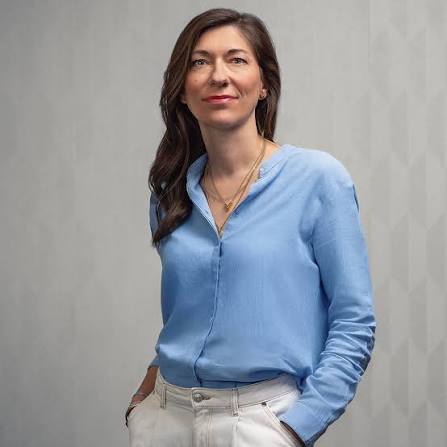 Une femme aux cheveux longs et noir. Elle est debout devant un fond neutre, elle porte un chemisier bleu clel.