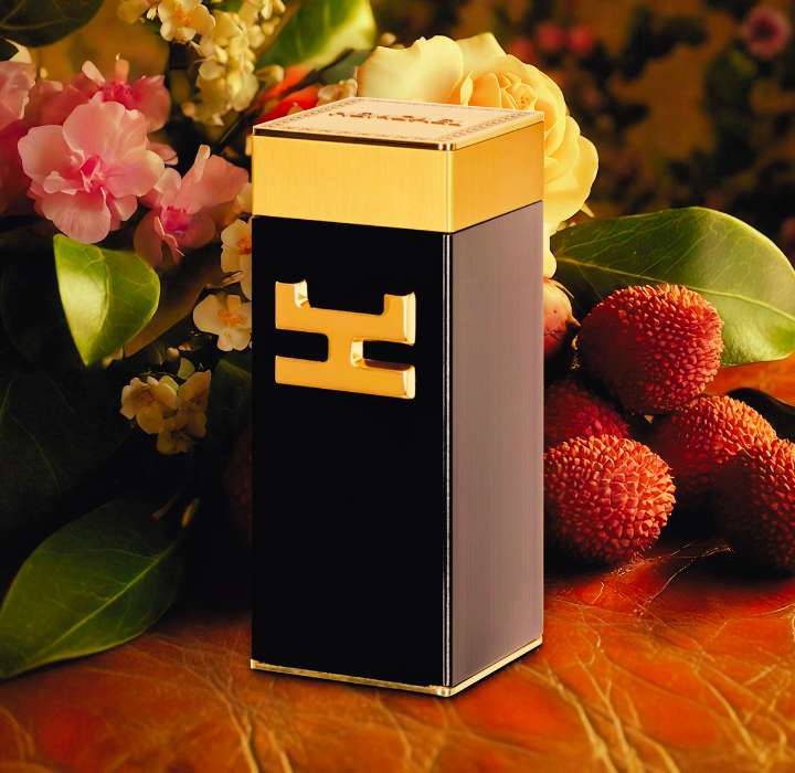 The image shows a bottle of Spiritus perfume by HEMCAEL. It sits on a leather surface, surrounded by colorful flowers and fruit. The bottle has a rectangular shape with a glossy black finish and a gold lid with an ornate border. On the front of the bottle, the gold HEMCAEL logo is visible in relief. In the background, delicate flowers in shades of pink and cream, and round fruit, likely lychees, add a touch of color and freshness to the composition. The soft lighting highlights the textures and details, creating a warm and refined ambiance. The overall effect evokes a blend of sophistication and nature, suggesting a rich sensory experience.