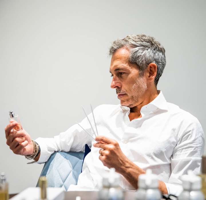 Un uomo con i capelli grigi, indossante una camicia bianca, sta esaminando attentamente un flacone di profumo in una mano, mentre con l'altra tiene dei bastoncini di test. È seduto a un tavolo, circondato da diversi flaconi di profumo.