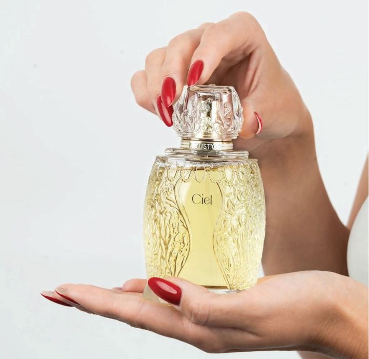 Eine weibliche Hand hält einen Parfümflakon aus bernsteinfarbenem Glas. Die andere Hand stützt ihn von unten. Die Fingernägel sind rot lackiert. Der goldene Verschluss ist kunstvoll geformt, und auf dem Etikett des Flakons steht „Ciel“.