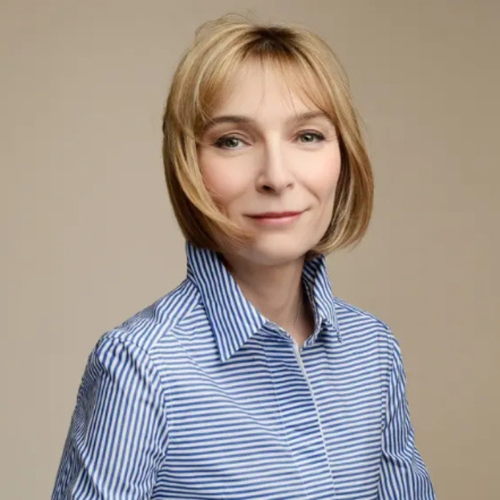Véronique Nyberg indossa una camicetta a righe blu e bianche. Sorride. Dietro di lei, uno sfondo neutro beige.