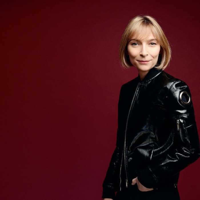 Véronique Nyberg è in piedi su uno sfondo rosso scuro. È di tre quarti e indossa un giubbotto di pelle nero.