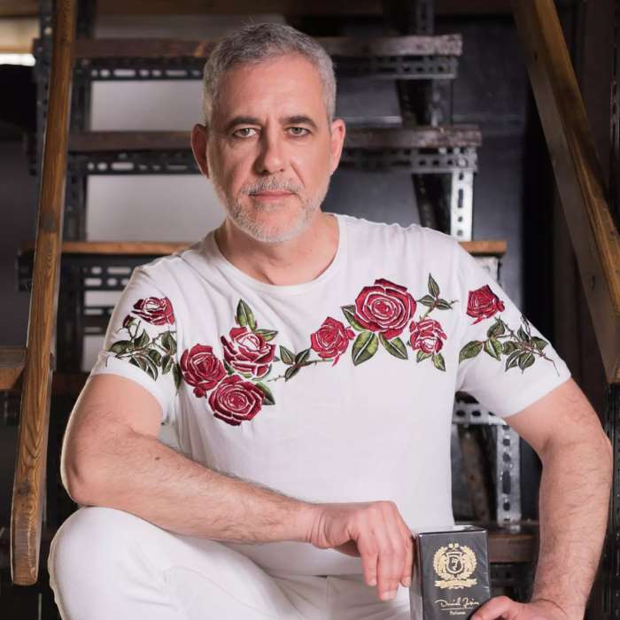 The image shows a perfume bottle from the Electimuss brand, highlighted by colorful lighting. The bottle has an elegant shape, with a red base and shiny gold cap. In the background, colored glass panels in shades of yellow, violet and red create a vibrant, reflective light effect. The luxurious design of the bottle, with a decorative medallion, is accentuated by the reflections of the colors, adding an artistic dimension to the composition.