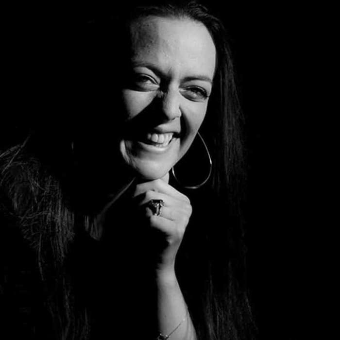 The image shows a smiling woman in dark lighting. Her face is partially lit, highlighting her bright smile and sparkling eyes. She has long hair, which falls freely around her face.

She wears large, eye-catching earrings and a visible bracelet on her wrist. Her hand is raised close to her face, with a friendly gesture.