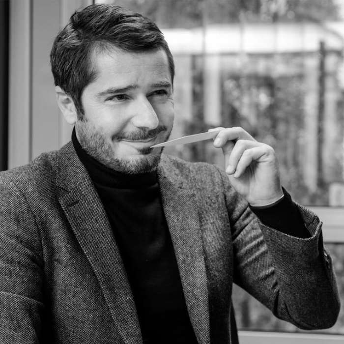 Black-and-white portrait of a man in a jacket and turtleneck, holding a smelling strip or a piece of paper near his face, with a blurred background.
