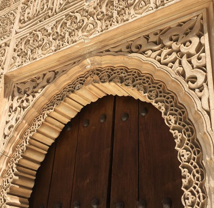L'image montre une porte en bois massif, ornée de détails architecturaux en pierre sculptée. La porte, de teinte sombre, est encadrée par un arc en pierre qui présente un travail de marqueterie délicat. L'arc est décoré de motifs géométriques et floraux complexes, typiques de l'architecture traditionnelle arabe. Les sculptures sont finement réalisées, mettant en valeur des volutes et des motifs entrelacés qui attirent le regard. Les bords de l'arc sont légèrement en relief, ajoutant de la profondeur et une dimension tridimensionnelle au design. Au centre, de gros clous métalliques sombres sont visibles sur la porte, soulignant son caractère robuste et ancien. La lumière éclaire la pierre, révélant des nuances de beige et de crème, tandis que les ombres accentuent les détails sculptés. L'ensemble évoque une atmosphère historique et artisanale, témoignage d'un savoir-faire traditionnel.