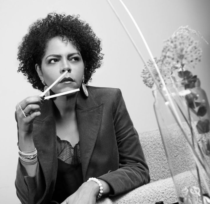 This black and white photo captures a portrait of Maiada El Khalifa, founder of the niche perfume brand TAYSHABA, sold on the SCENTISSIME website. We see a woman with a confident, creative look. Her curly hair frames her expressive face as she holds a pencil pensively, suggesting she might be an artist or designer in her field. Her elegant yet casual blazer, paired with discreet jewellery, reflects a sophisticated yet accessible style. Her determined gaze and confident posture exude a strong, inspiring presence. The blurred background emphasises her silhouette while revealing organic elements such as plants, adding a natural touch to the image. The lighting highlights her expressive features, capturing her creative intensity. Overall, this portrait reveals a talented and passionate woman, who seems to draw her inspiration from nature to create something unique and authentic, whether in art, design or niche perfumery.