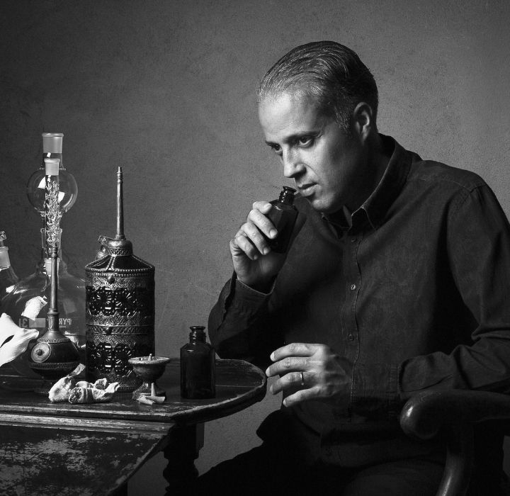 L’image montre le fondateur de MAISON INCENS, Philippe Constantin, assis à une table, examinant divers flacons et objets disposés devant lui. Il sent le contenu de l’un des flacons, évaluant probablement les parfums. La table est ornée de carafes ornées, de flacons et d’autres verres, ce qui suggère qu’il s’engage dans une sorte d’appréciation ou de critique de niveau professionnel ou connaisseur. La scène en noir et blanc donne à l’image une atmosphère artistique et pensive alors que l’homme se concentre intensément sur sa tâche, les sourcils froncés. Dans l’ensemble, la composition et le cadre suggèrent qu’il s’agit d’un artisan qualifié ou d’un expert examinant son métier avec un œil et un nez avertis. La marque MAISON INCENS est vendue sur la boutique en ligne SCENTISSIME.