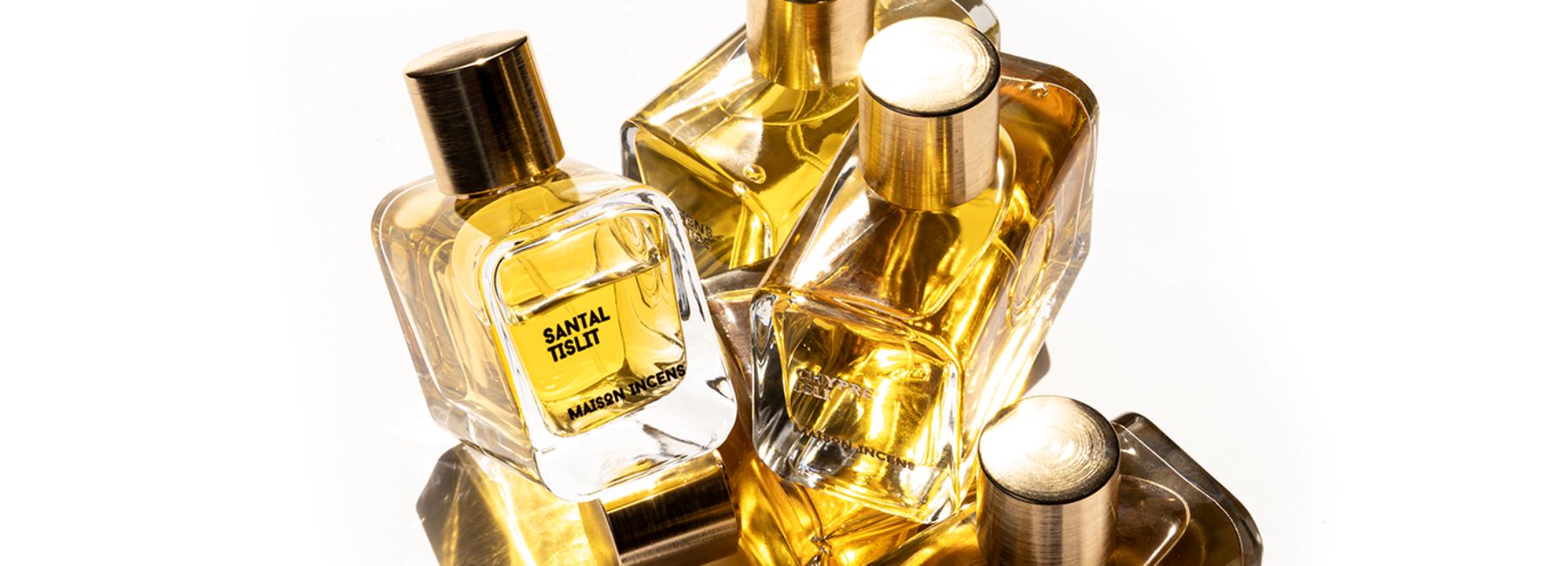 L’image présente trois flacons de parfums de niche, chacun contenant un liquide doré. Les bouteilles sont disposées en diagonale du bas à gauche vers le haut à droite sur une surface réfléchissante. Le flacon au premier plan est étiqueté « SANTAL TISLIT » et « MAISON INCENS » et possède un bouchon métallique. La deuxième bouteille est partiellement cachée par la première, tandis que la troisième bouteille est montrée de dos. La marque MAISON INCENS est vendue sur la boutique en ligne SCENTISSIME.