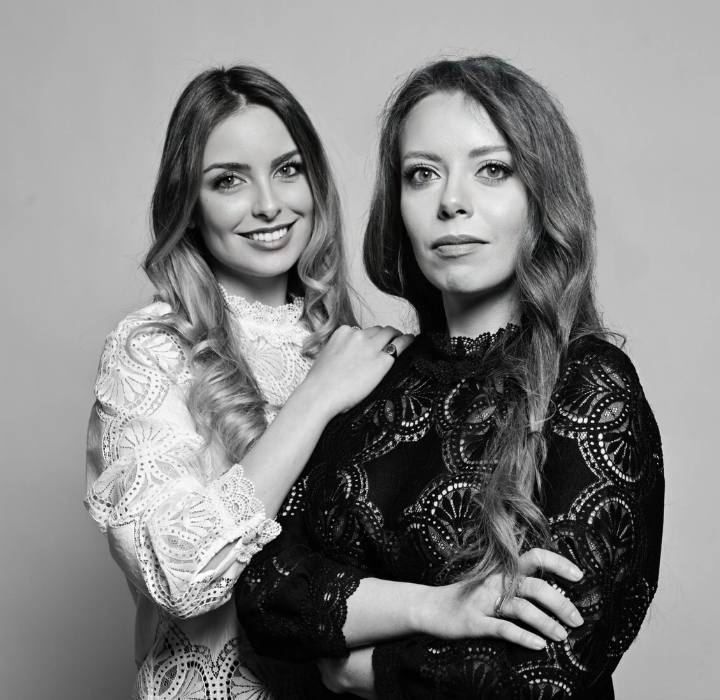 L'image en noir et blanc présente deux femmes posant ensemble. La femme à gauche est la parfumeure Sofia Bardelli. Elle a de longs cheveux blonds et porte une blouse blanche ornée de motifs délicats. Elle sourit chaleureusement.La femme à droite, Fabiola Bardelli, a des cheveux longs et châtains, elle porte une blouse noire également décorée de motifs subtils. Son expression est plus sérieuse et réfléchie.Elles se tiennent l'une à côté de l'autre, la femme de gauche ayant légèrement sa main posée sur l'épaule de la femme de droite, ce qui souligne une connexion entre elles. L'arrière-plan est simple et neutre, ce qui permet de mettre en valeur les figures et les vêtements des deux femmes.