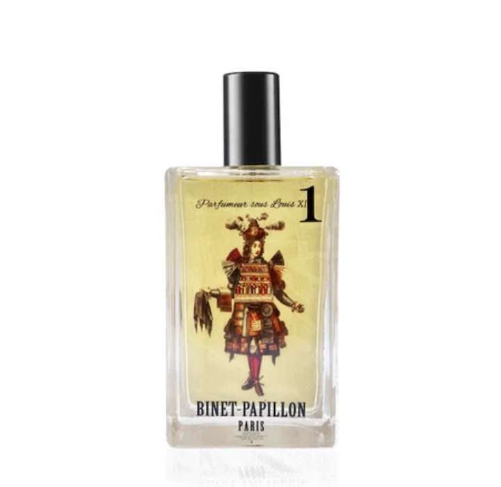 The image shows a rectangular perfume bottle with a black cap. The label depicts an illustration of a 16th or 17th century perfumer, dressed in a colorful and ornate manner, with elements resembling royal attire. The background of the label is pale yellow, and the name