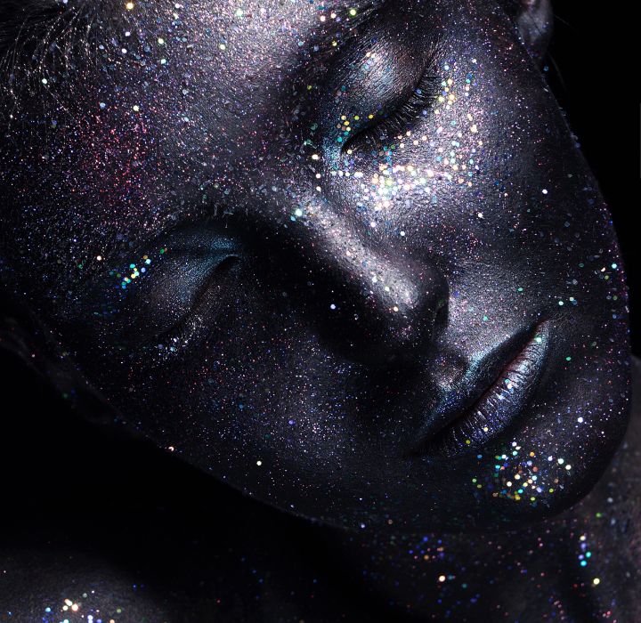 L’image montre le visage d’une personne avec un maquillage qui ressemble à une galaxie ou à une nébuleuse. Le maquillage est principalement noir et scintillant, couvrant la zone autour des yeux, du nez et des joues. Il présente un dégradé de couleurs, allant de nuances plus foncées au centre à des nuances plus claires avec des étincelles multicolores près des bords, créant l’illusion d’un ciel nocturne rempli d’étoiles et de poussière cosmique. Cette photo illustre le concept de la marque de parfum de niche MAISON INCENS, vendue sur la boutique en ligne SCENTISSIME.