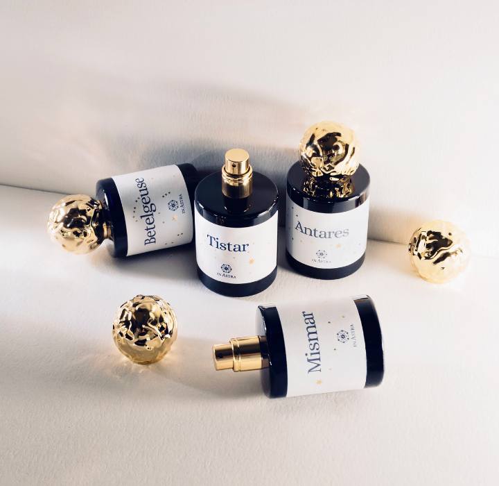 L'image montre quatre flacons de parfum disposés artistiquement sur une surface claire, créant une ambiance raffinée. Chaque flacon a un design similaire, avec un corps en verre noir et une étiquette blanche sur laquelle des noms sont inscrits en police élégante. Les noms des parfums sont